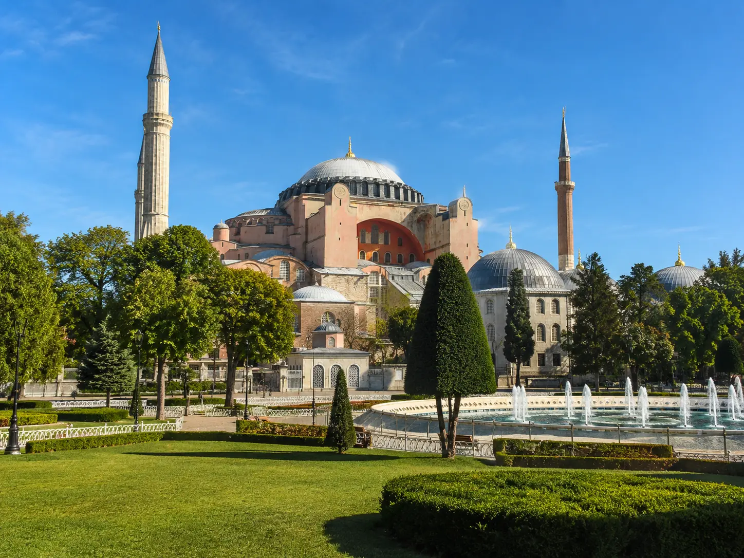 Hagia Sophia seen from a landscaped garden on a clear sunny day, with fountains, trees, domes, and minarets visible from a shifted park perspective.