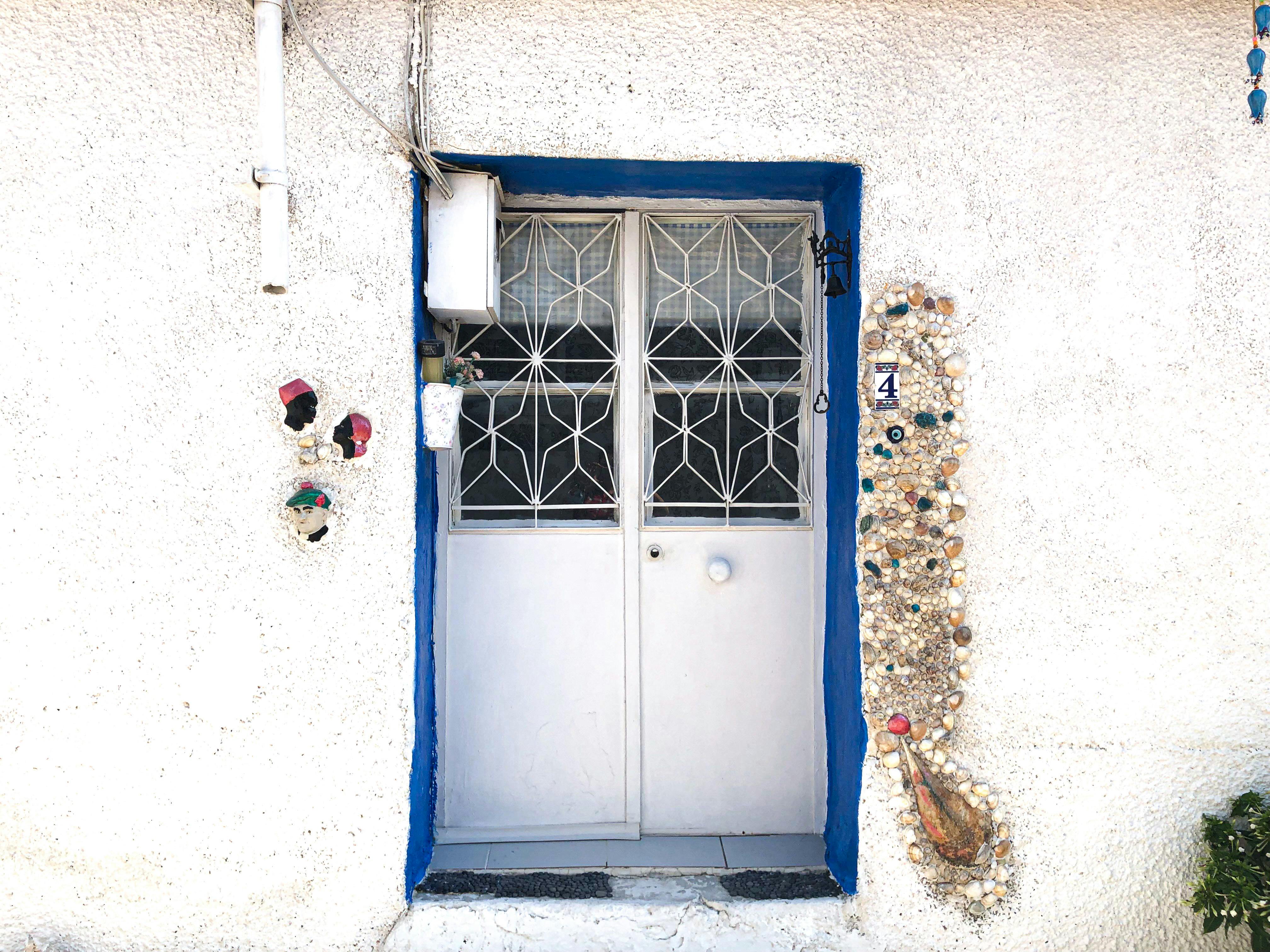 A white wall in Ayvalık, Balıkesir, Türkiye, featuring arts and crafts elements with a blue-framed door accent.
