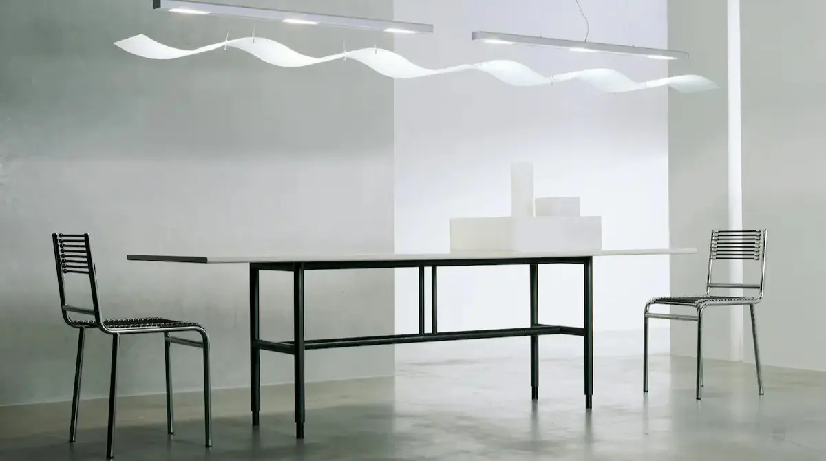 A minimalist modern dining setup with a rectangular glass-top table, black metal base, and several chairs near the wall in a space with mostly white tones.