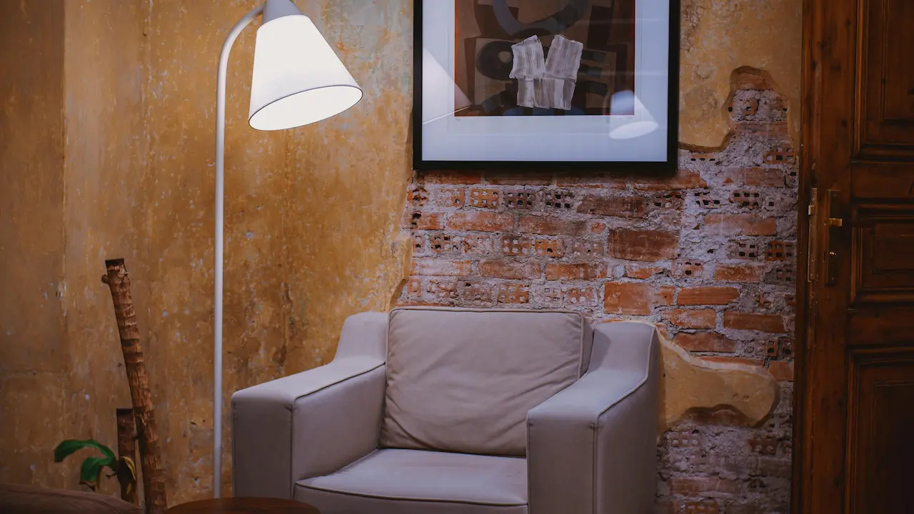 Rustic stone wall with a white armchair next to a standing lamp in the room.