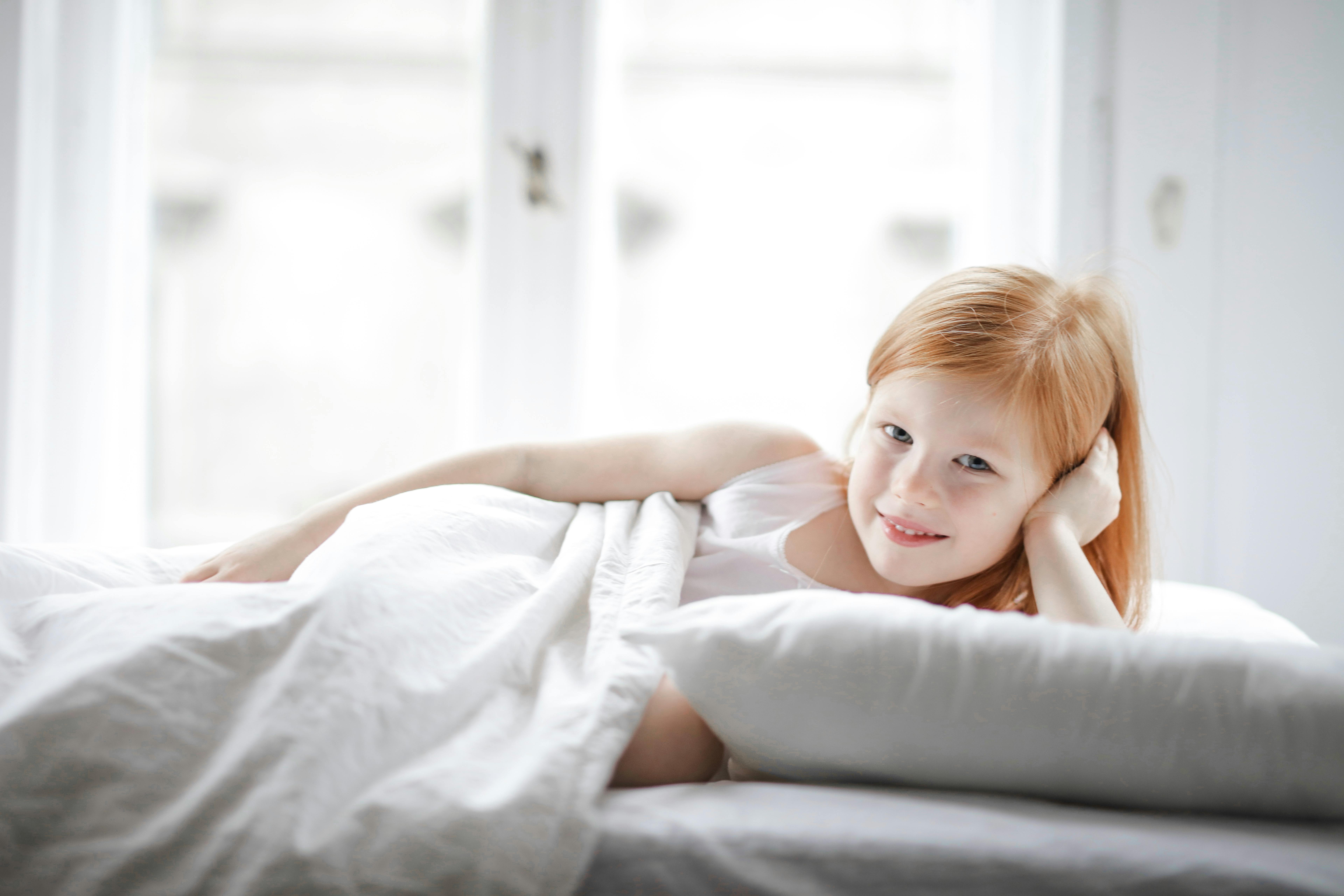 Girl lying on white bed in naturally lit room with soft, bright colors, creating a peaceful, uplifting mood.