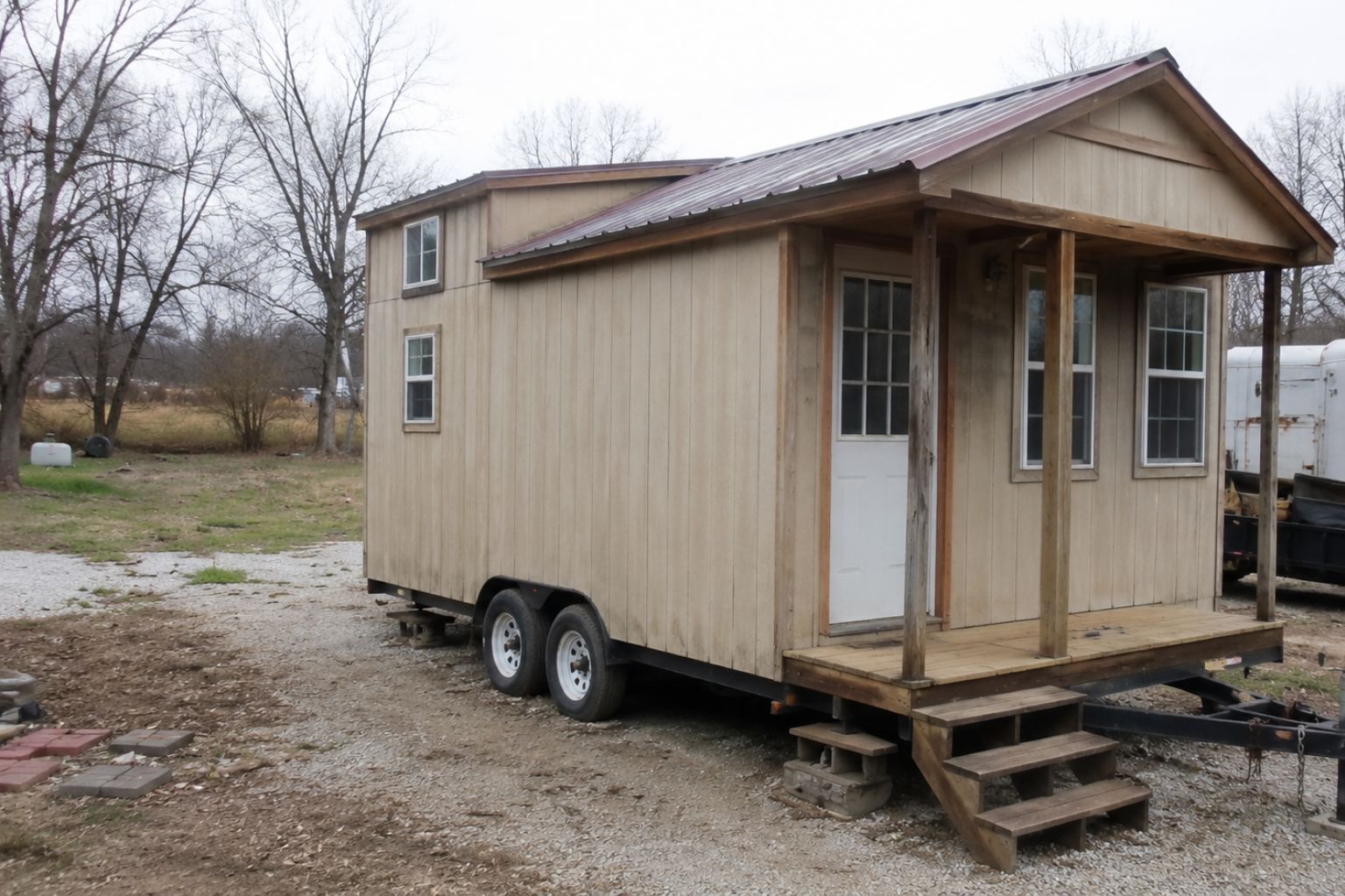 Budget tiny house on wheels with simple siding, metal roof, small porch, trailer frame, and gravel yard.