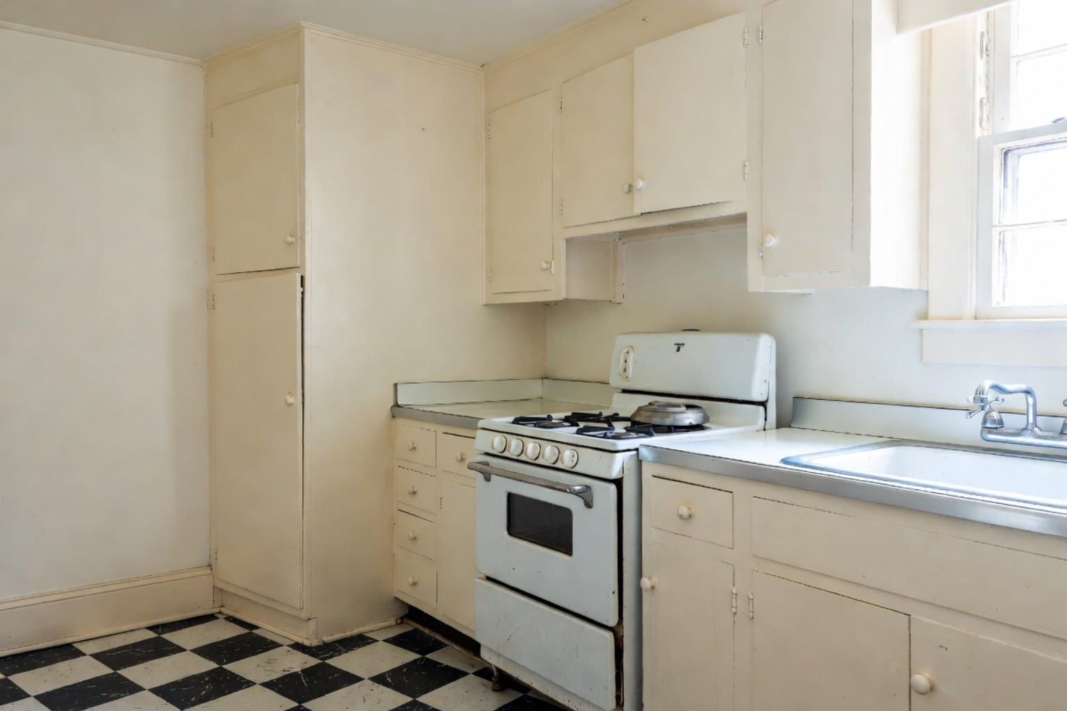 1930s-style kitchen with cream cabinets, white stove, checkerboard floor, and simple sink.