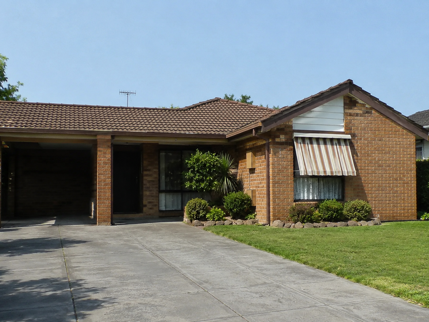 Close-up view of a modest 1980s brick suburban house with a low roofline, recessed entry, front driveway, awning, and foundation planting.
