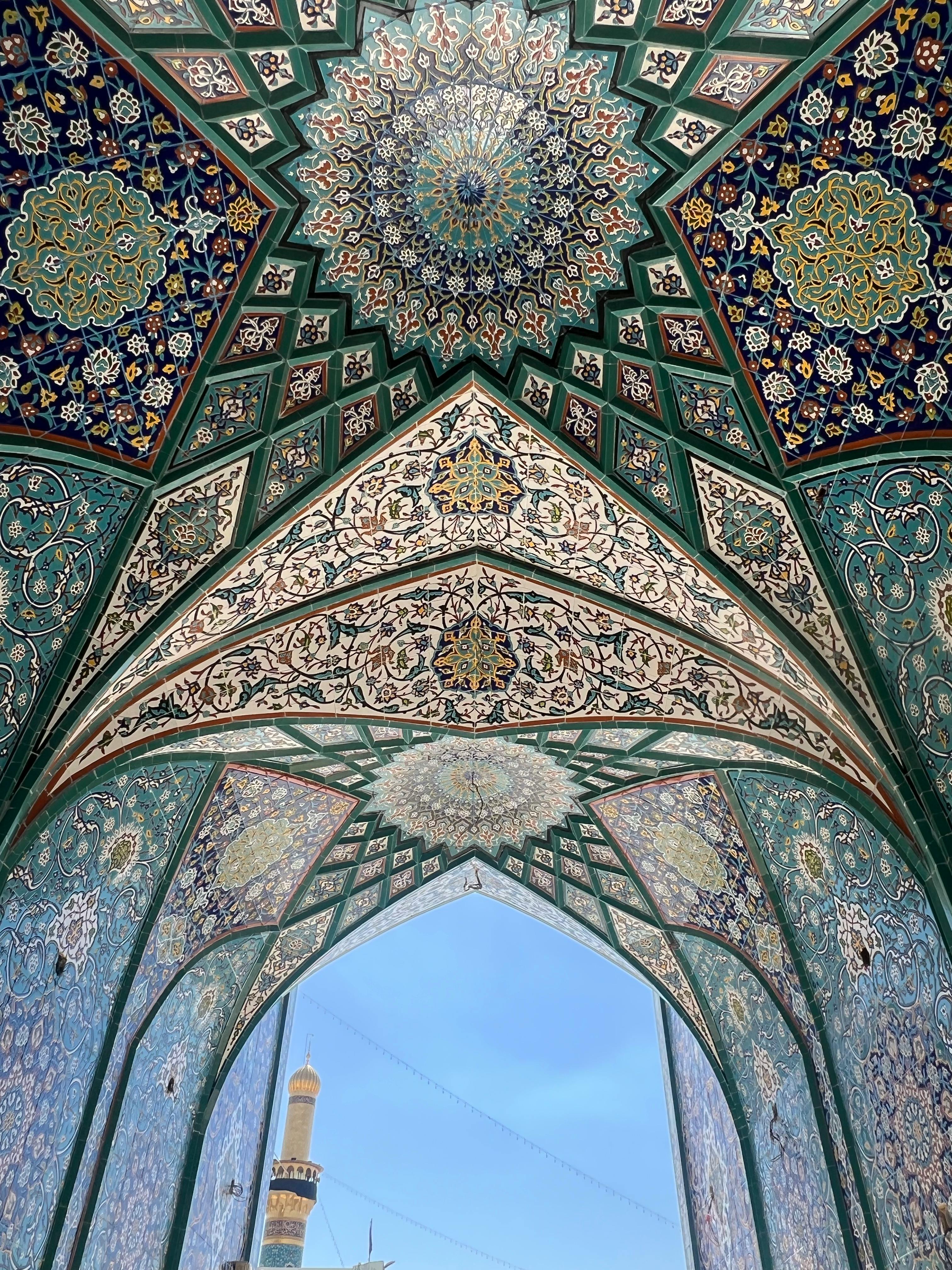 Intricate Persian muqarnas ceiling with floral tilework and blue sky framed through arched gateway
