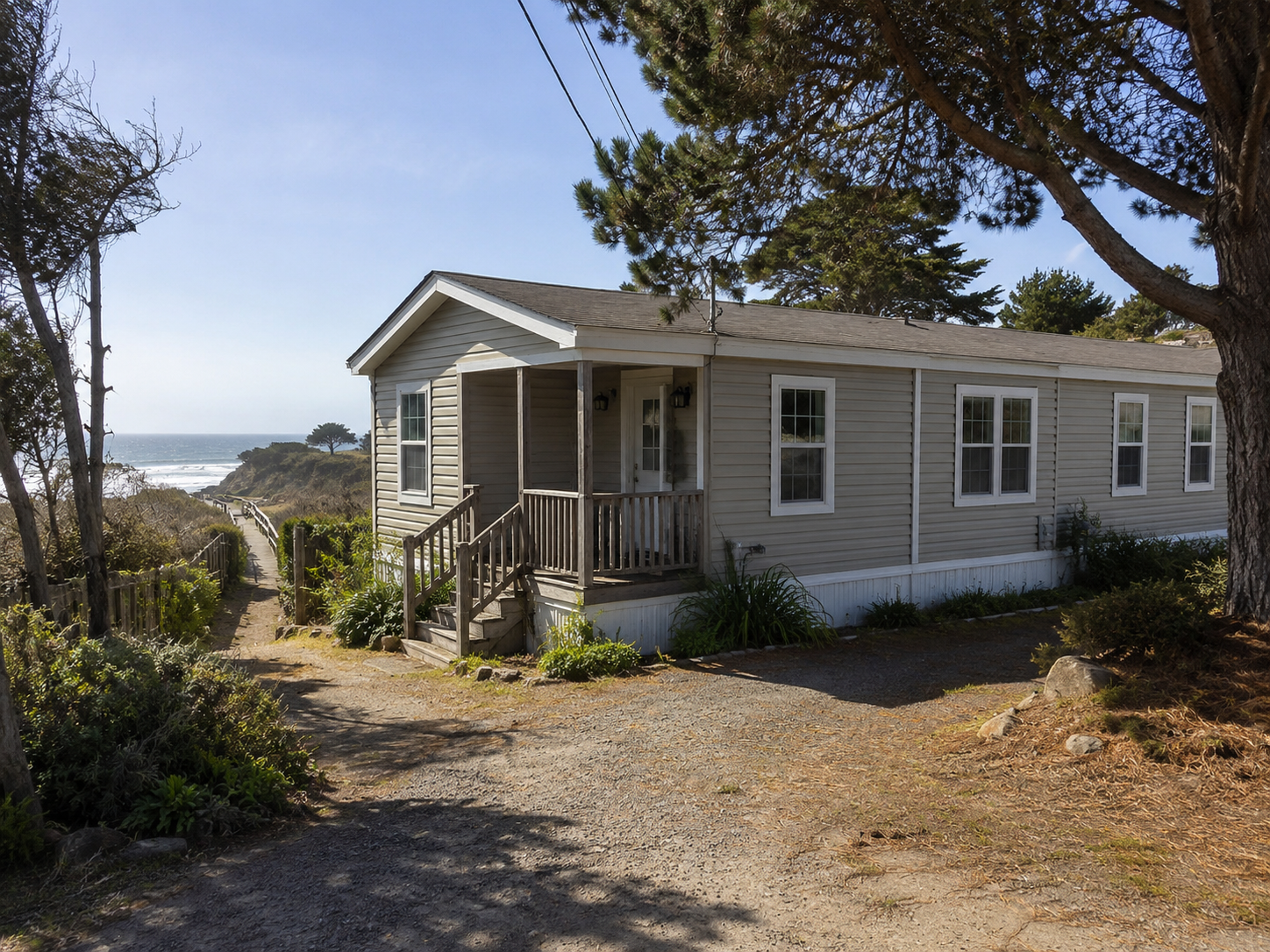 Single-wide manufactured home near a coastal bluff with gravel drive, porch, trees, and path leading toward the beach.