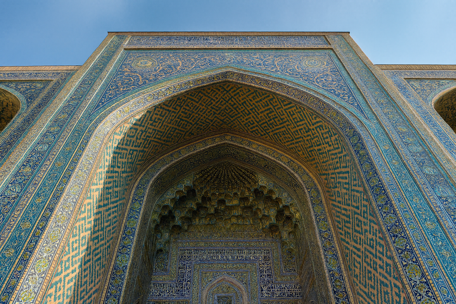 Upward view of an Islamic archway with muqarnas vaulting and blue geometric tile patterns.
