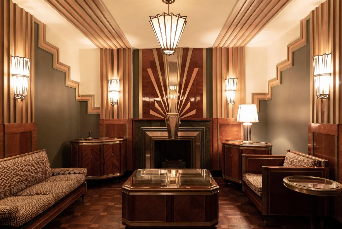 Art Deco sitting room with stepped wall geometry, polished wood paneling, symmetrical lighting, and a sunburst fireplace design.