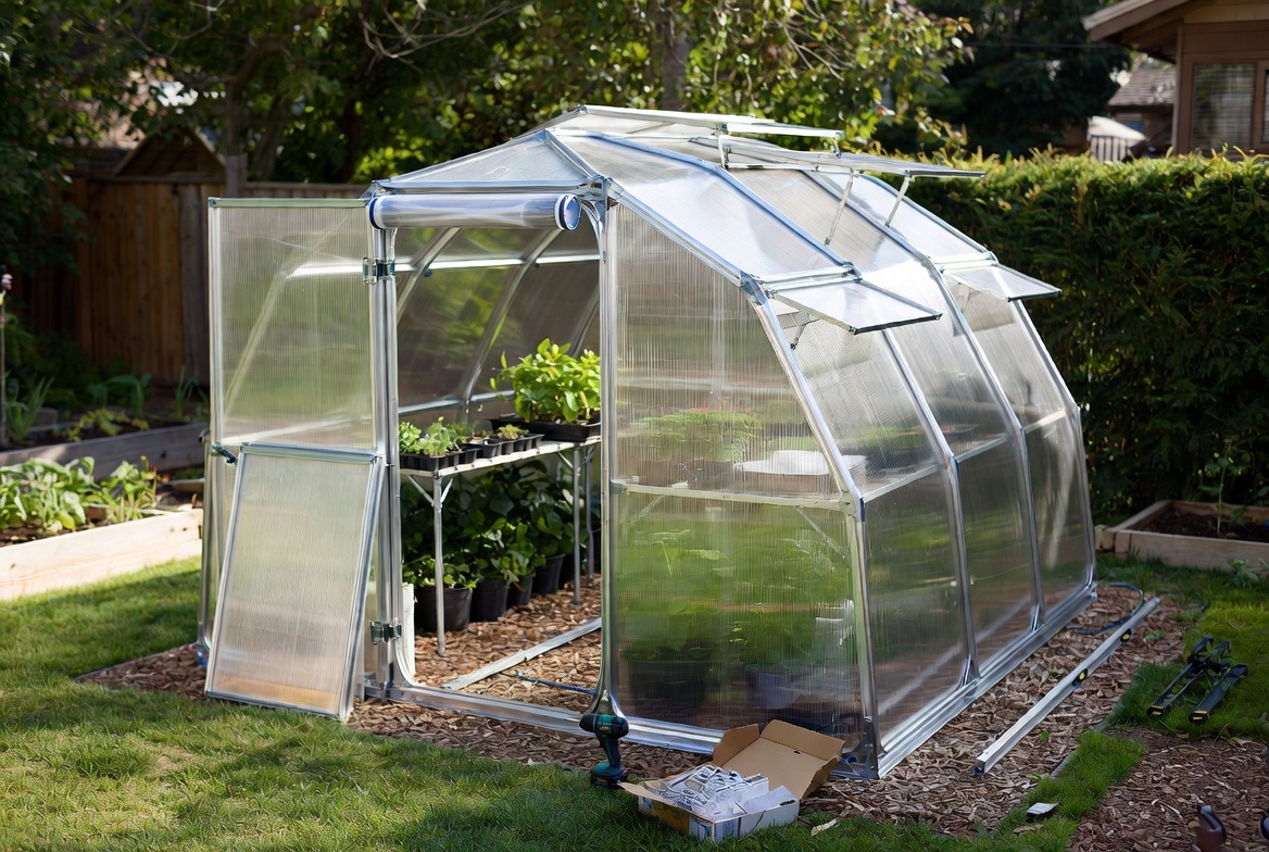 Small polycarbonate backyard greenhouse kit with open door, roof vents, and visible assembly hardware.