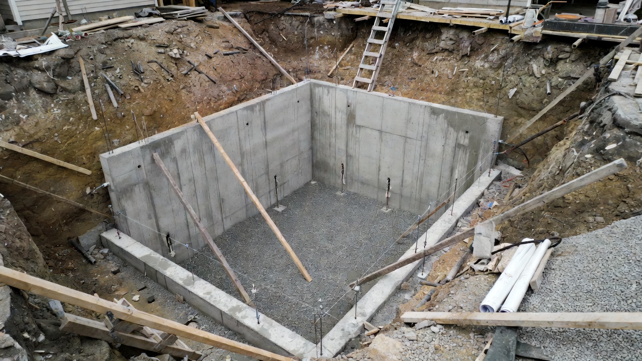 Basement foundation under construction with poured concrete walls, gravel base, and deep excavation.