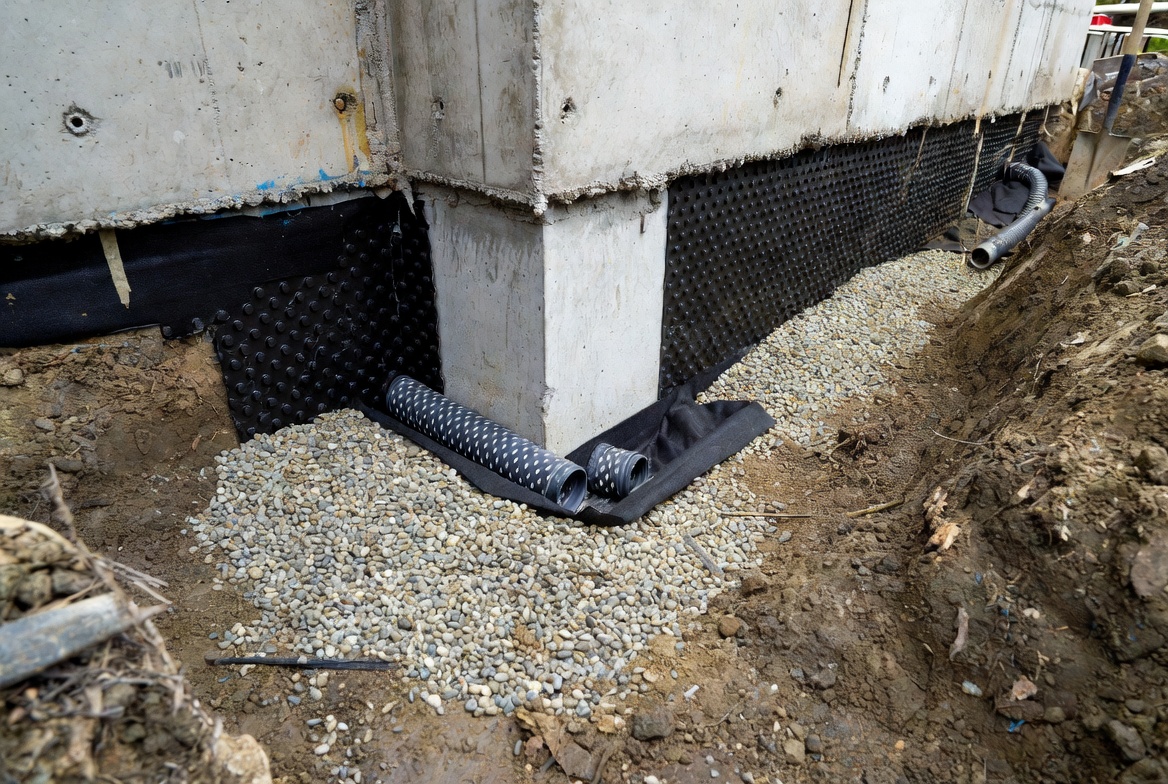 Exterior foundation footing drain with gravel, pipe, and drainage board at the wall base.