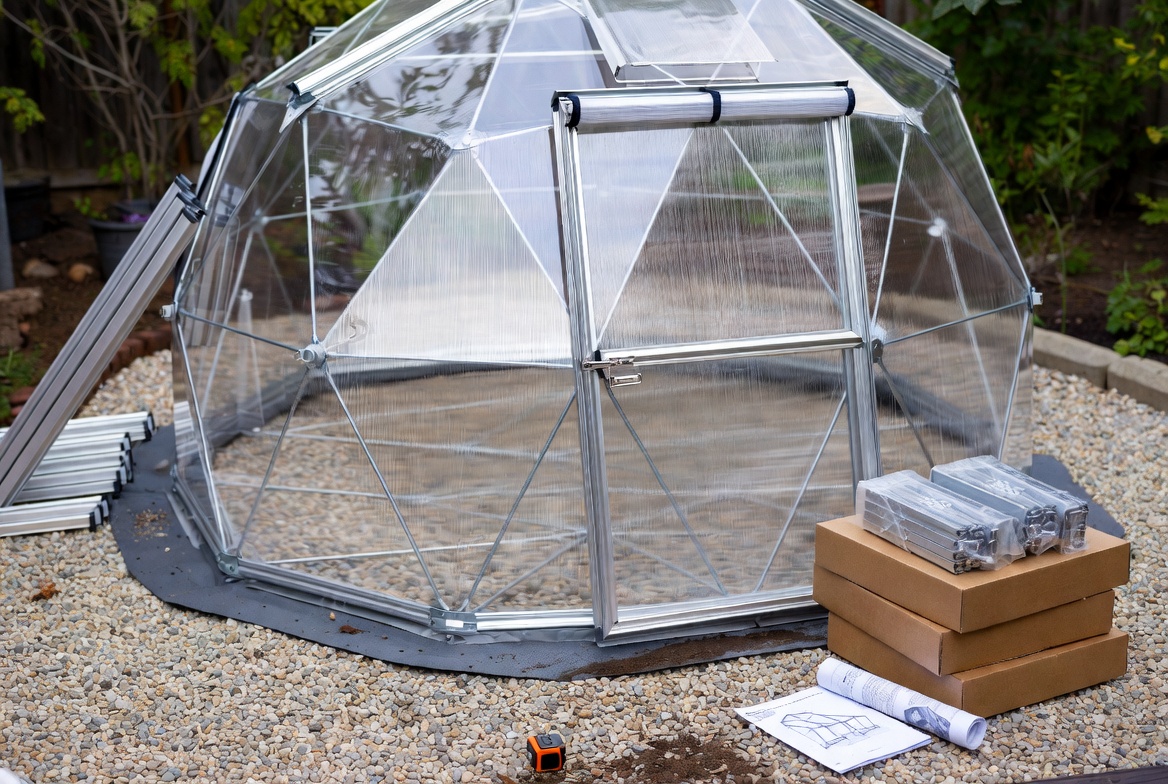 Greenhouse dome kit with visible frame, polycarbonate panels, door, and boxed components on a gravel base.
