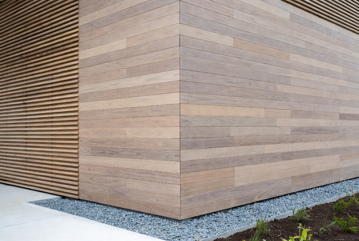 Modern facade corner with horizontal timber cladding, slatted timber screen, and gravel drainage strip.