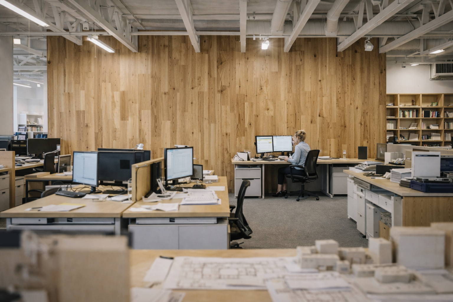 Female architect working alone in a modern open-plan office with wood wall and desks.
