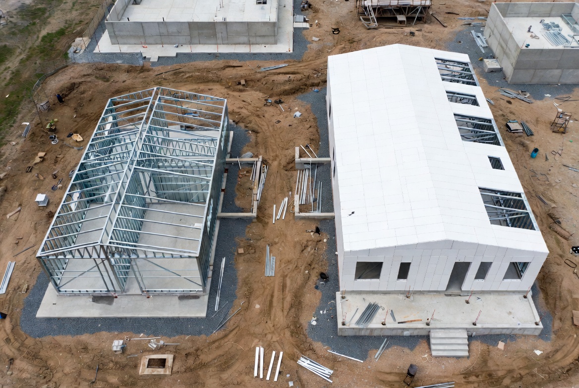 Prefab metal building shell beside a finished wall-panel structure on a construction site.