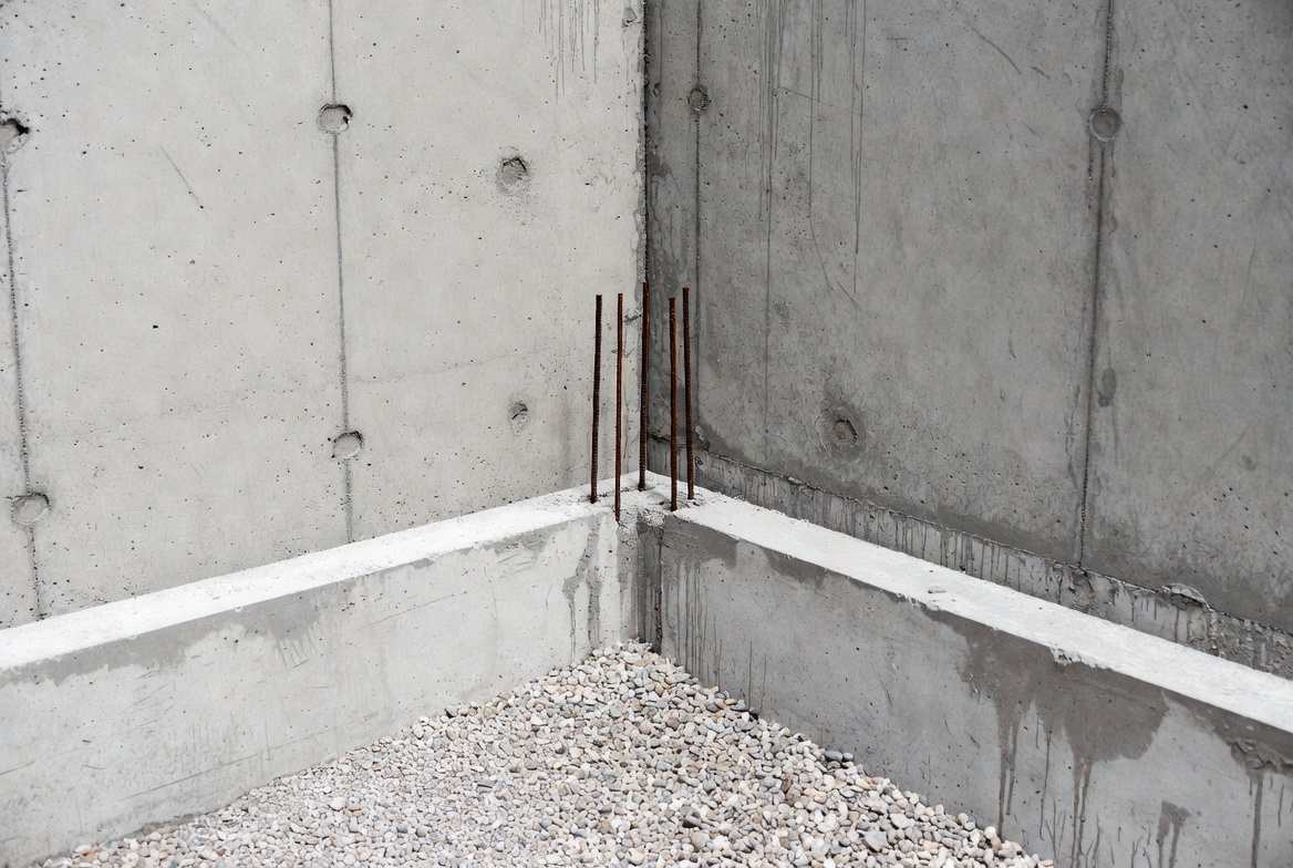 Reinforced concrete foundation corner with footing ledge, gravel base, and exposed rebar.