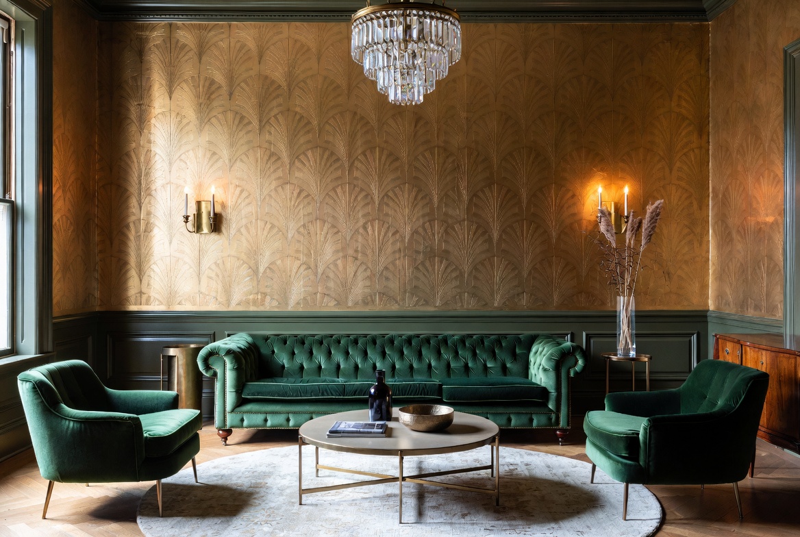 Authentic 1920s Art Deco living room featuring an emerald green velvet Chesterfield sofa, metallic gold fan-pattern wallpaper, crystal cascade chandelier, and matching velvet armchairs.