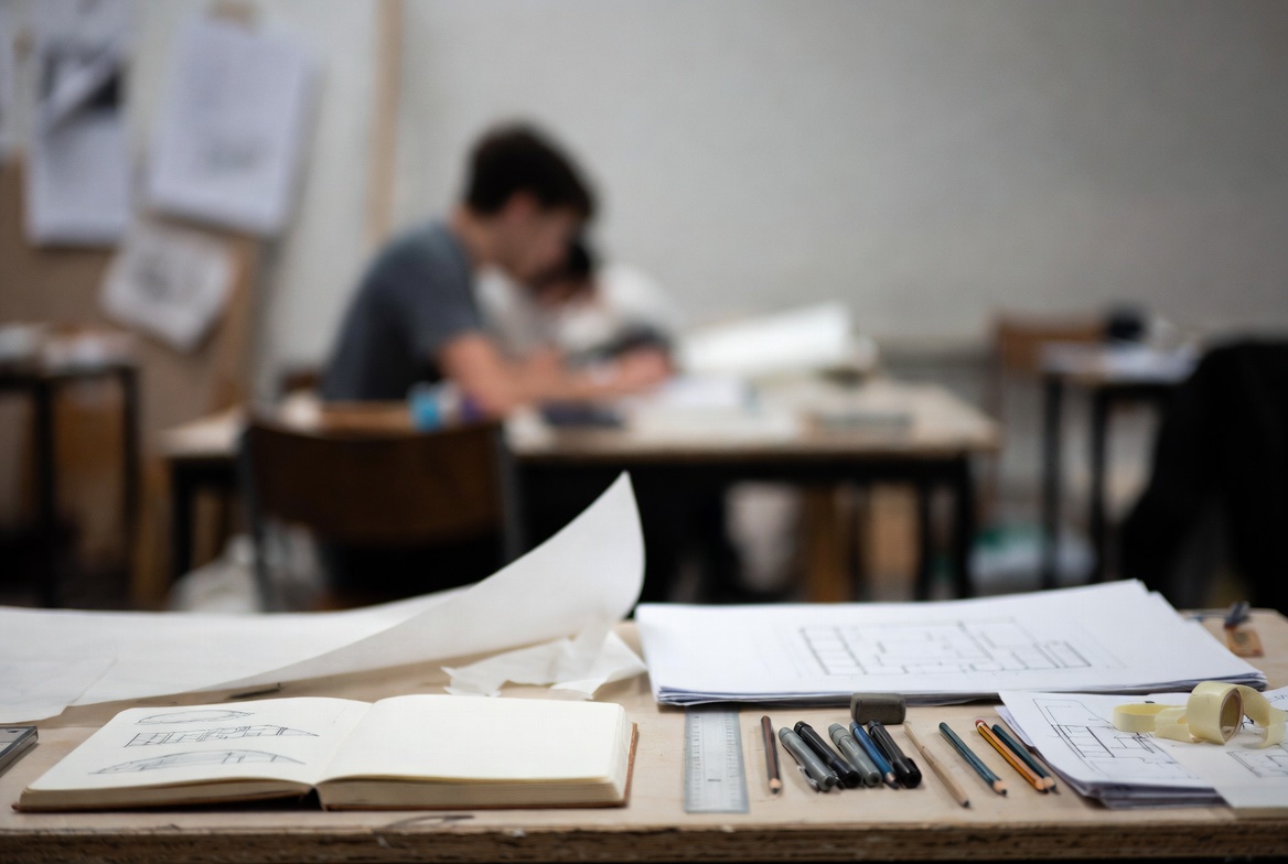 Architectural drawing tools, sketchbook, and plan sheets on a studio desk with students working in the background.