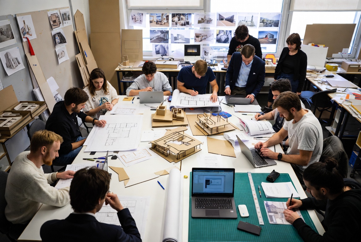 Architecture students working around a studio table with drawings, models, laptops, and project materials.