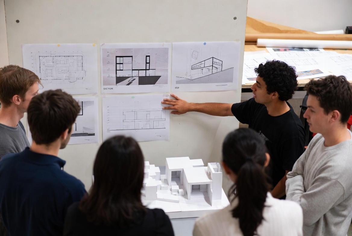 Architecture students reviewing pinned drawings and a study model during a studio critique.