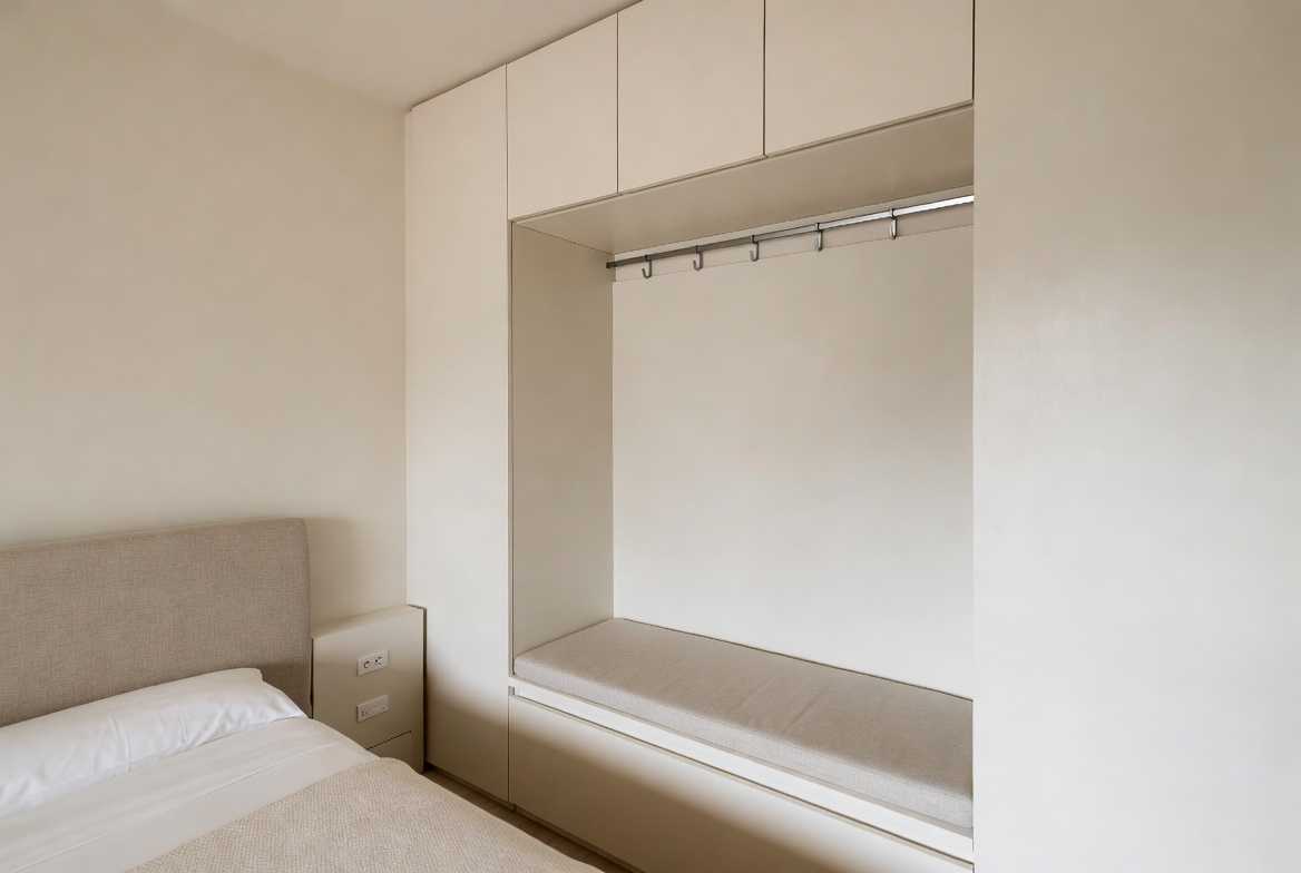 Small bedroom with built-in bench niche, overhead cabinets, and integrated wall storage beside the bed.