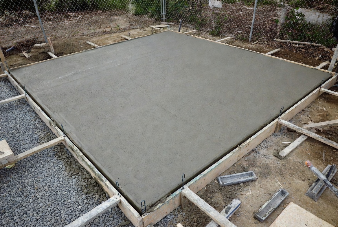 Fresh concrete pad inside wood formwork with gravel base visible around the slab edges.