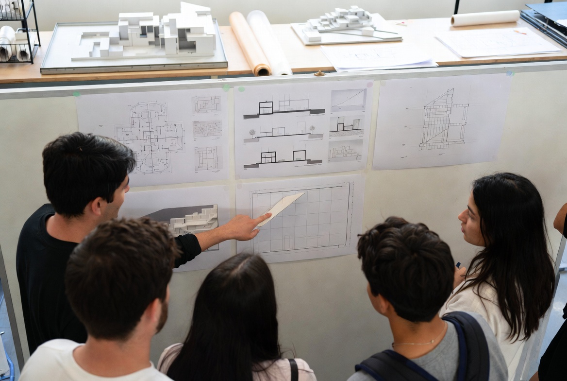 Architecture students reviewing pinned plans, sections, and a study model during a studio discussion.
