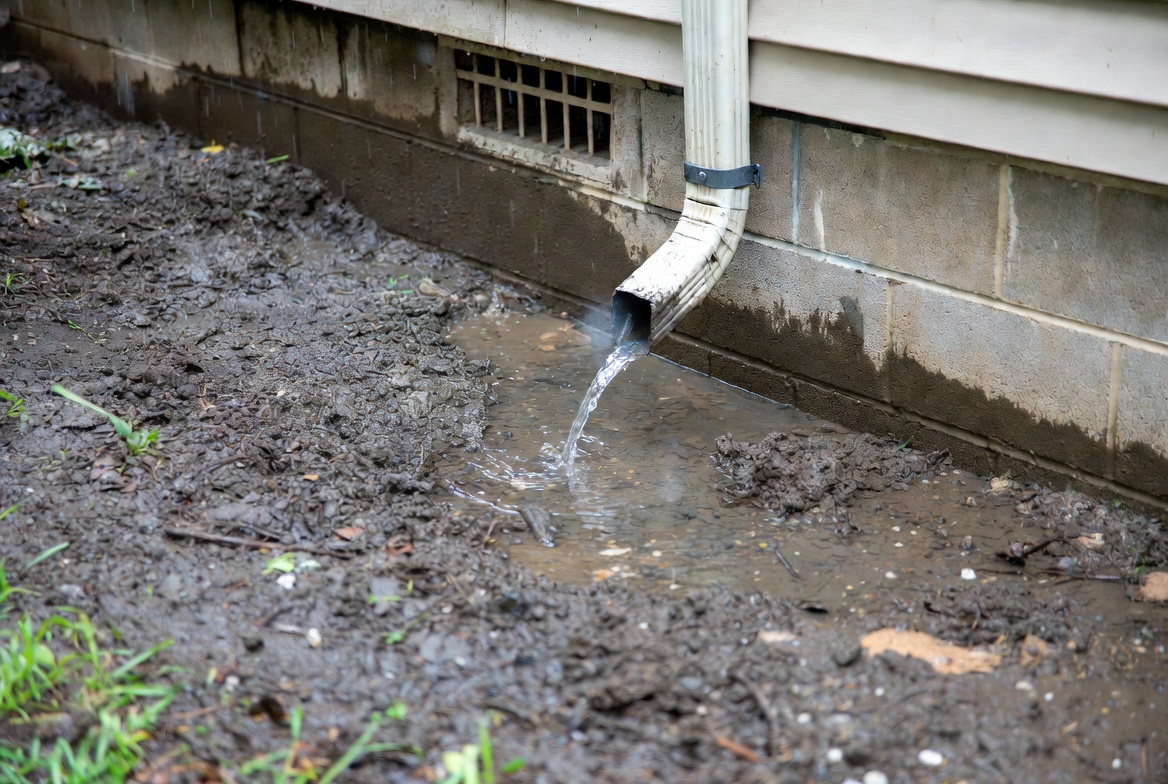 Downspout discharging beside a crawl vent along a block foundation wall.