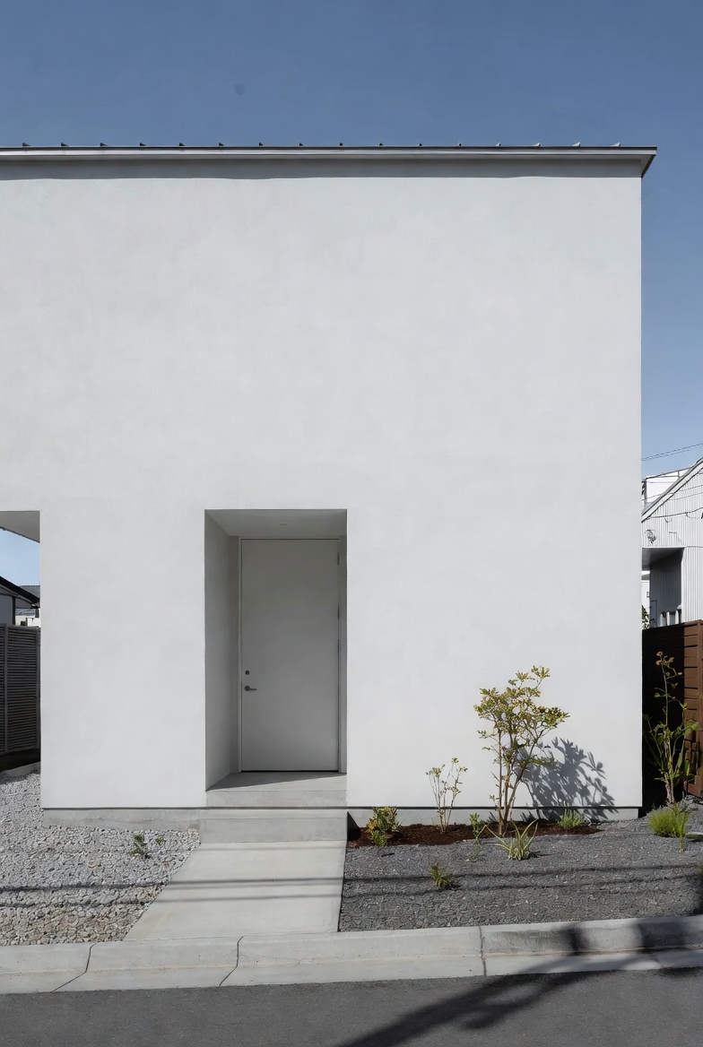 Minimal small-house front with a simple entry path, plain white facade, and restrained planting.