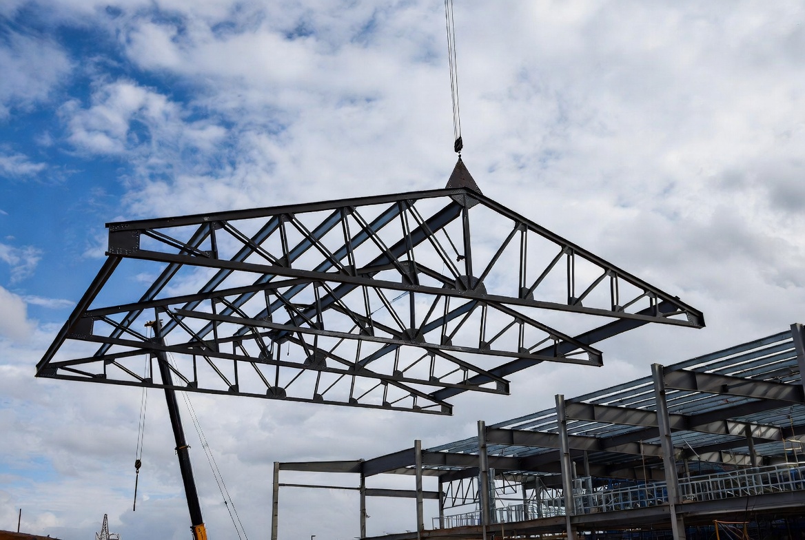 Steel roof truss being lifted by crane over a steel building frame.