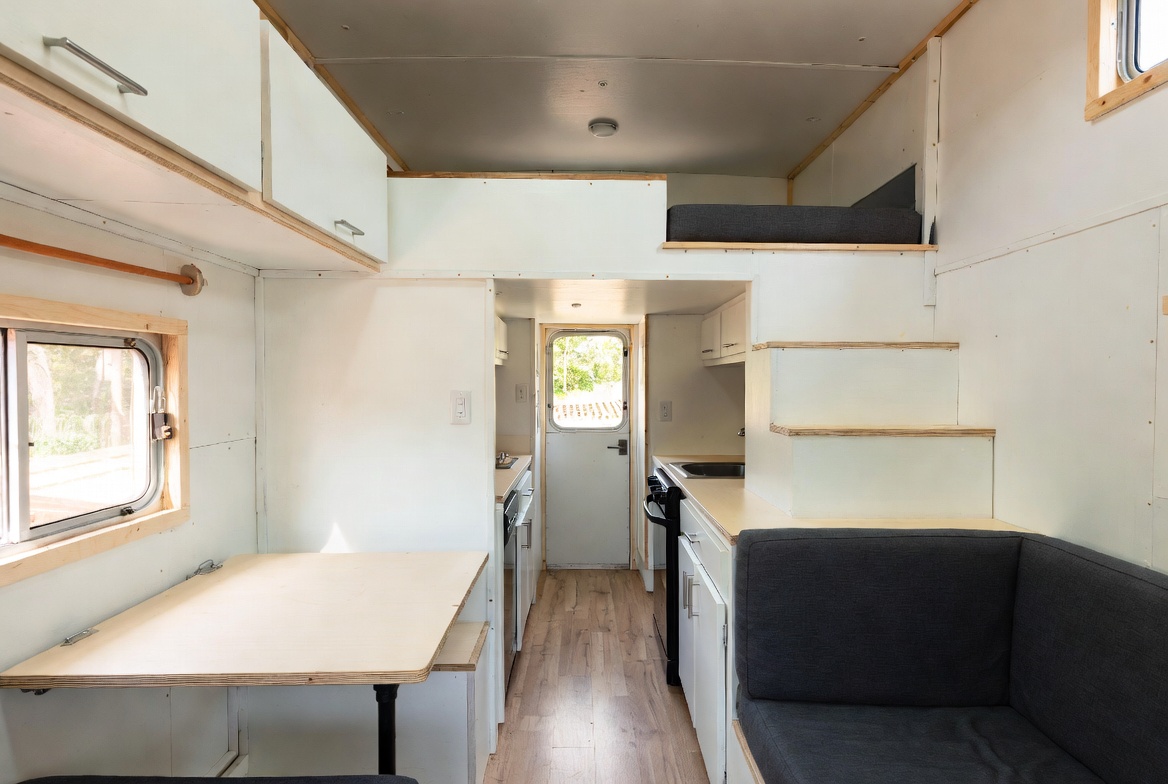 Tiny home interior with loft bed, built-in seating, table, and compact kitchen.
