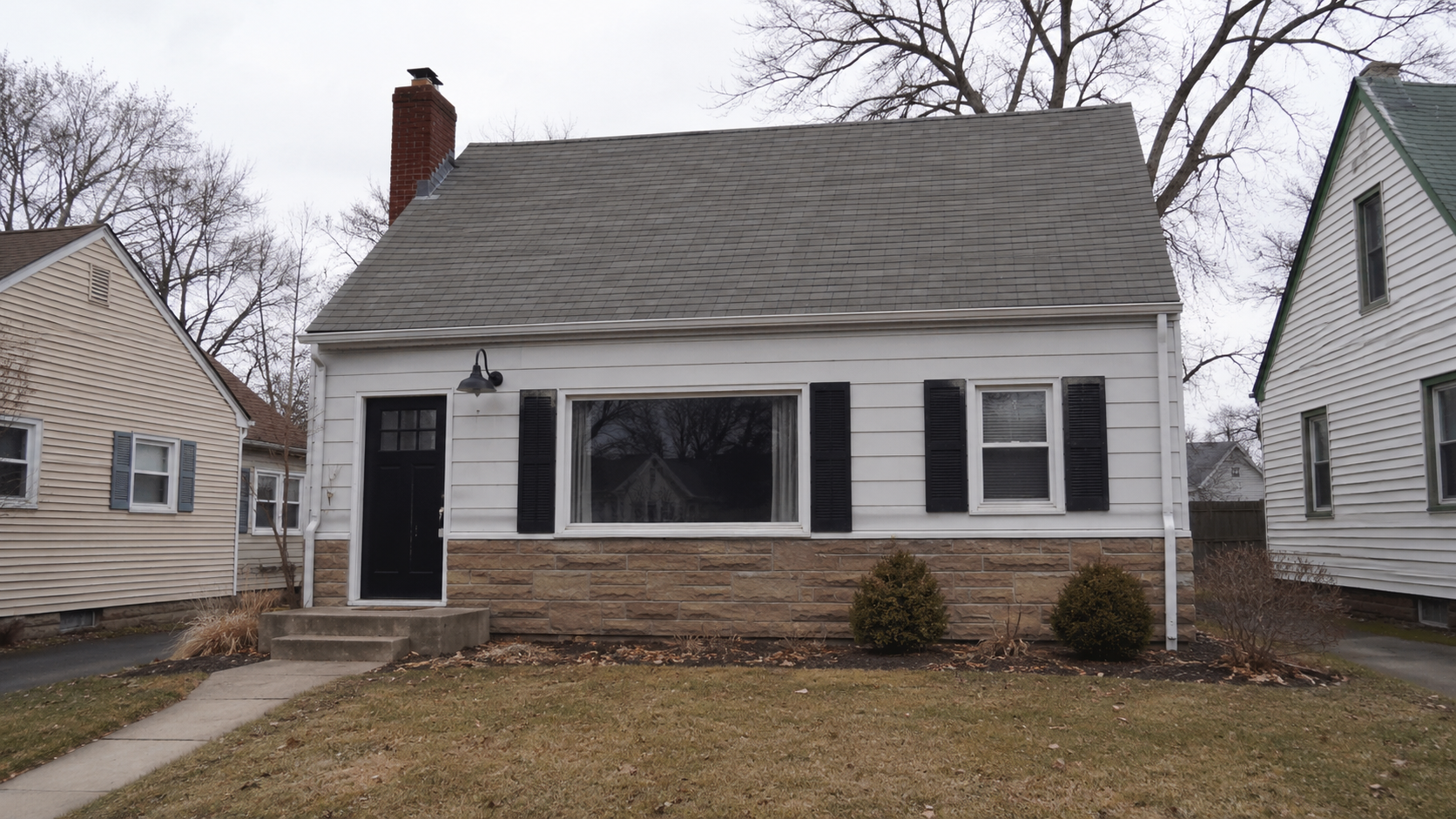Modest 1940s Minimal Traditional house with oversized picture window, fake stone veneer, black shutters, and mismatched exterior updates.