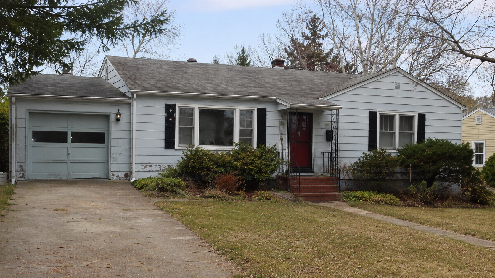 A modest 1950s postwar house before remodeling with an attached garage, low roofline, tired siding, black shutters, dated entry rails, and overgrown planting.