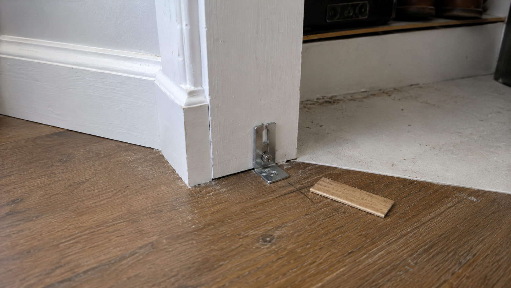 Close-up of a bifold door lower jamb bracket at the bottom of a closet opening, with finished flooring, baseboard trim, pencil layout marks, and a small wood shim nearby.