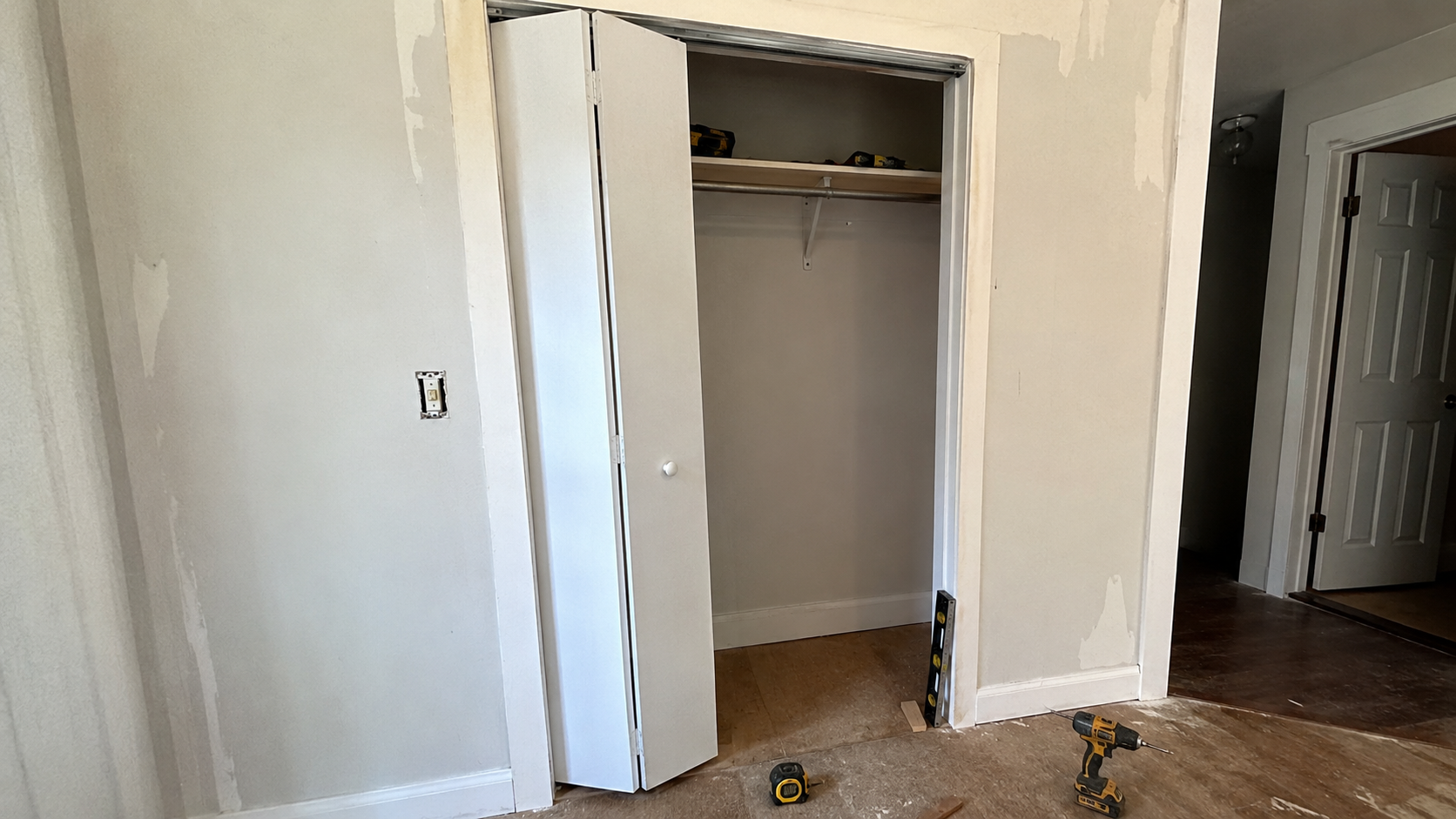 Partly installed white bifold closet door in an older home with the top track visible, tools on the floor, and a level and shim used to check the door opening.