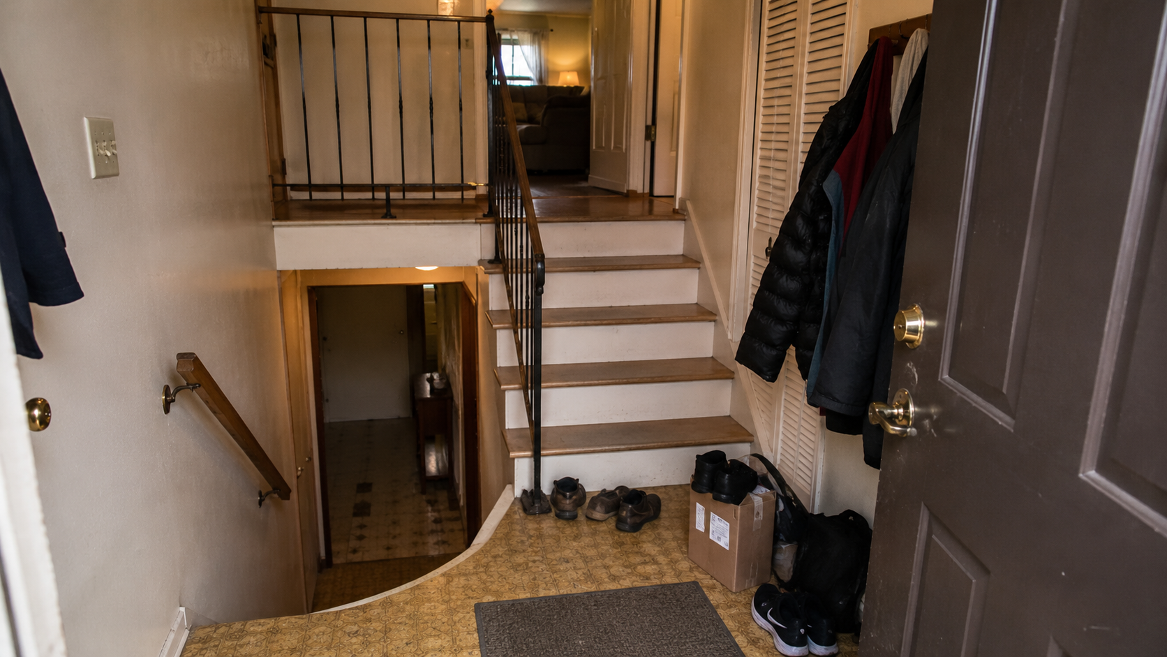 Cramped raised ranch split-entry landing with stairs up and down, coats, shoes, package, dated railing, and weak light.