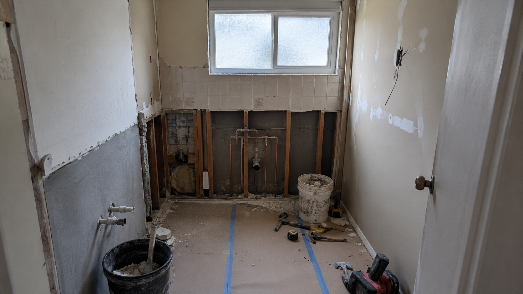 Older ranch bathroom mid-remodel with exposed wall framing, rough plumbing, old tile, patched drywall, tools, and protected flooring.