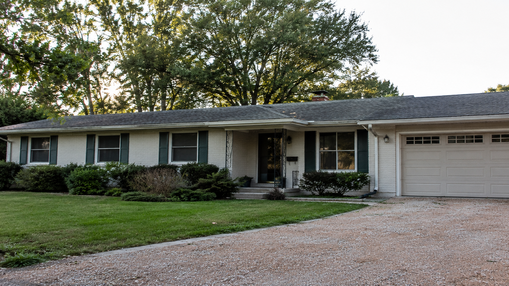 Older ranch house exterior with a low roofline, attached garage, plain entry, and aging landscaping.