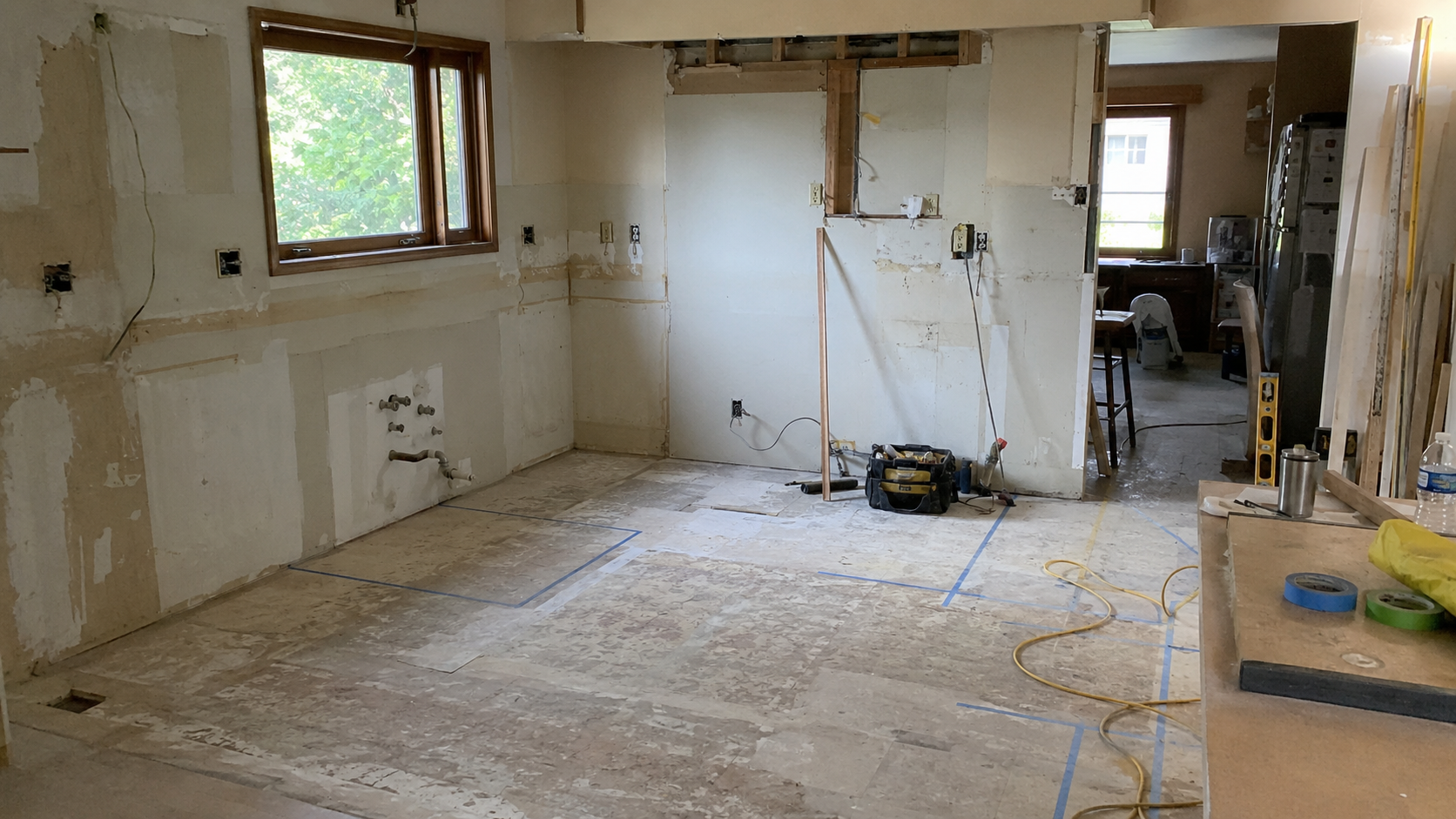 Ranch kitchen demolition showing stripped walls, old plumbing rough-ins, exposed wiring, floor layout markings, and an unfinished opening during a remodel.