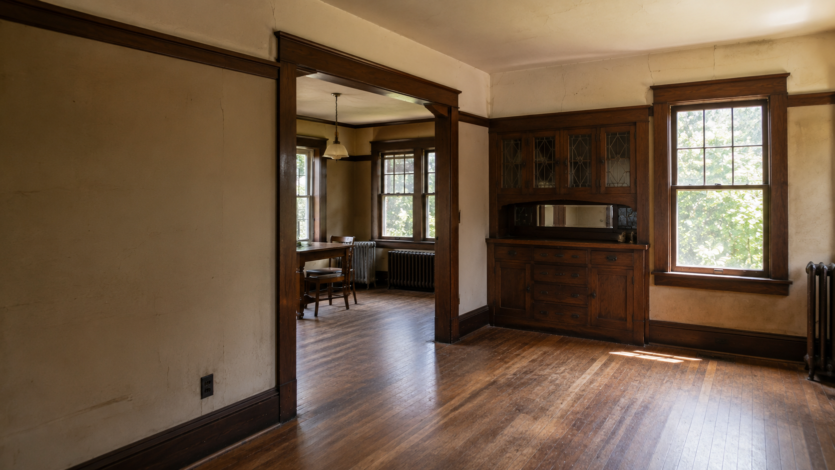 Original 1920s Craftsman interior with dark woodwork, built-in cabinet, radiator, and worn floors.