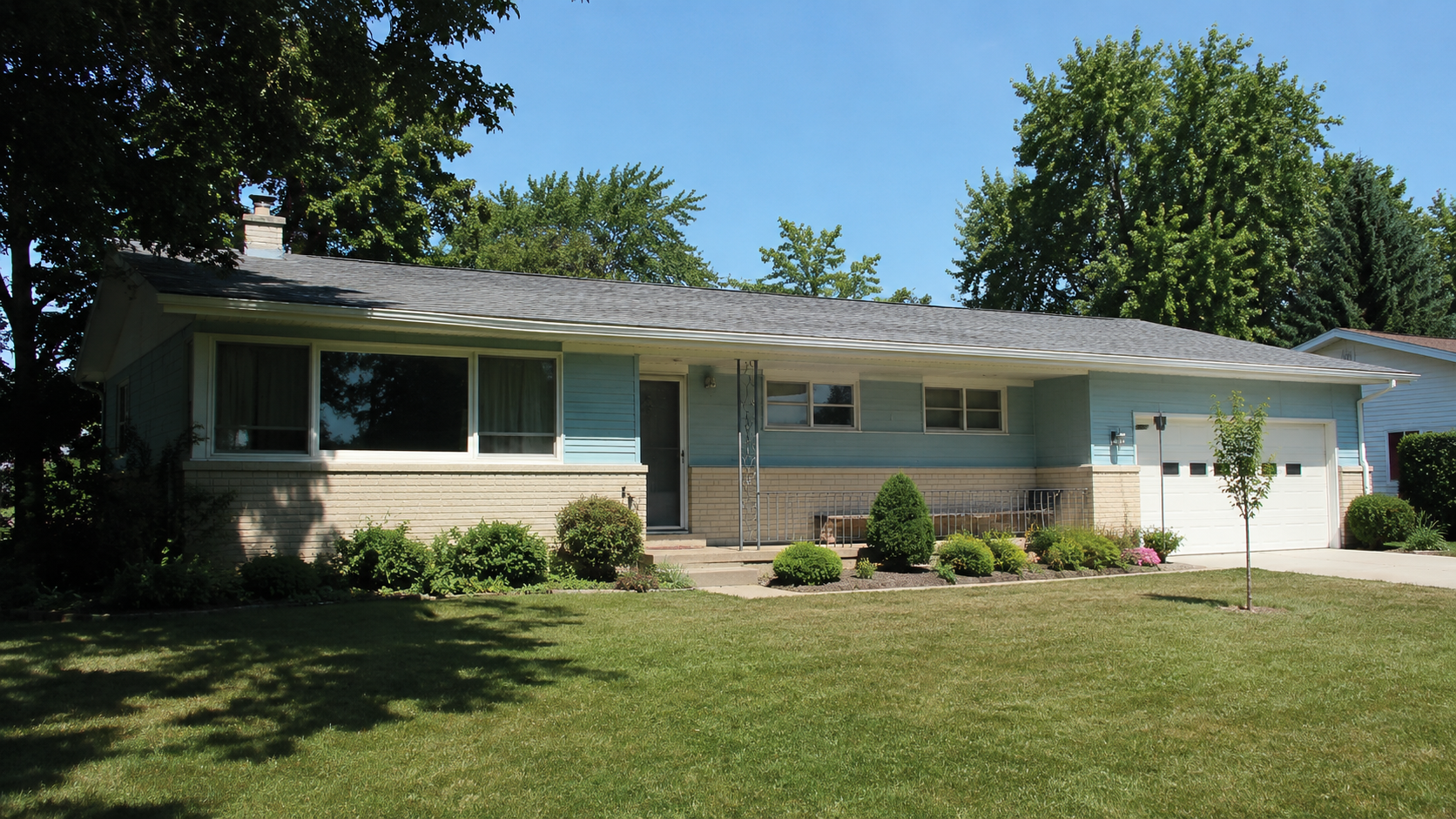 Modest 1950s ranch house with pale siding, brick base, low roofline, lawn, and attached garage.