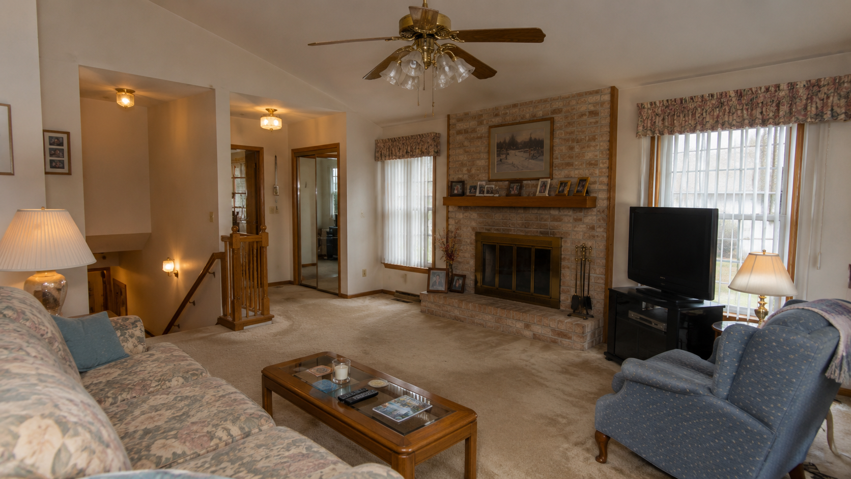 Dated 1980s family room with beige carpet, oak trim, brick fireplace, brass fan, vertical blinds, and mirrored doors.