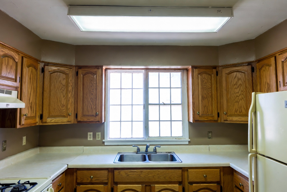 1980s kitchen sink wall with oak cabinets, laminate counters, and fluorescent ceiling light.