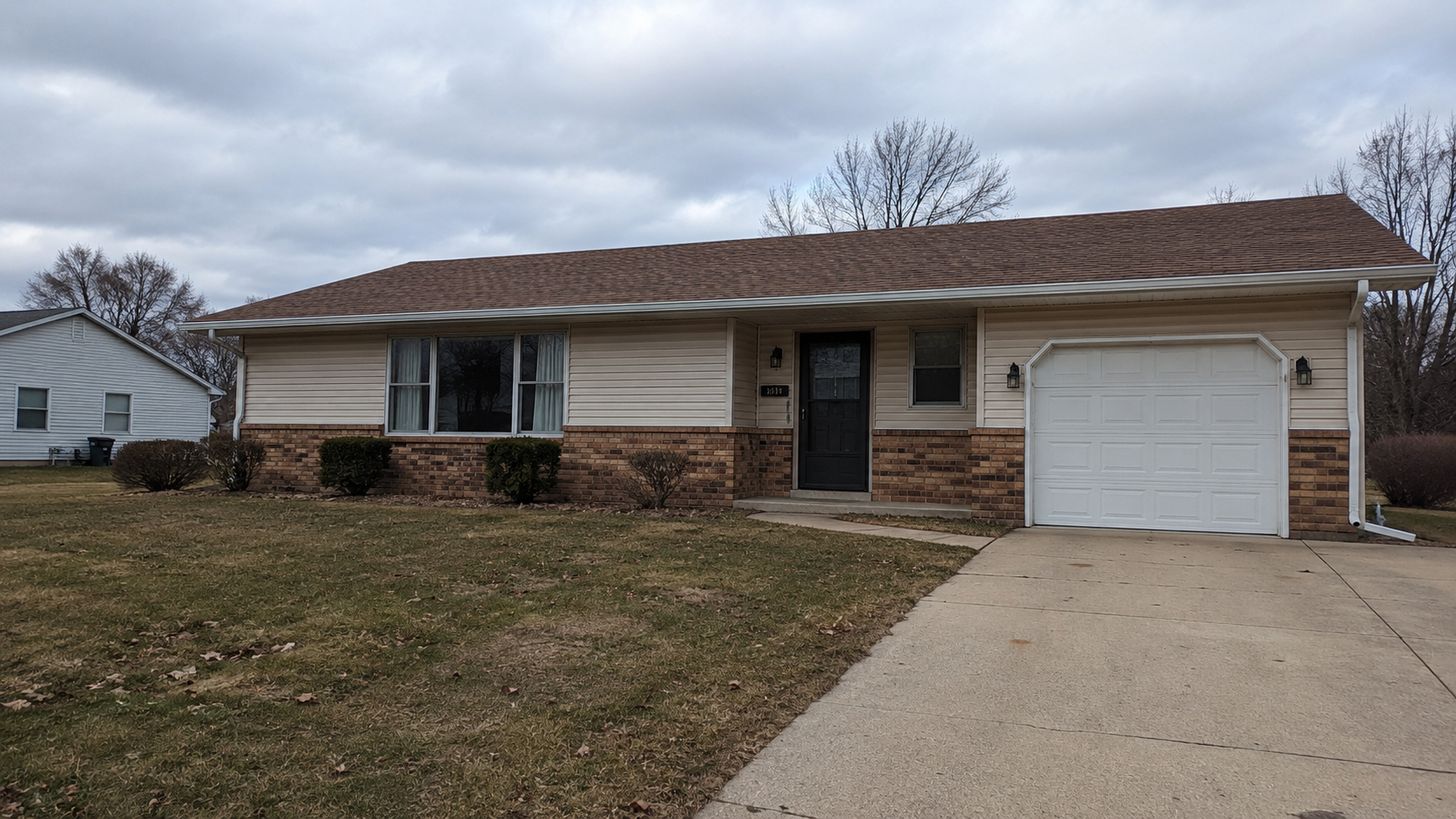 Modest 1980s suburban ranch house with beige siding, brown brick, front garage, storm door, low shrubs, and patchy lawn.