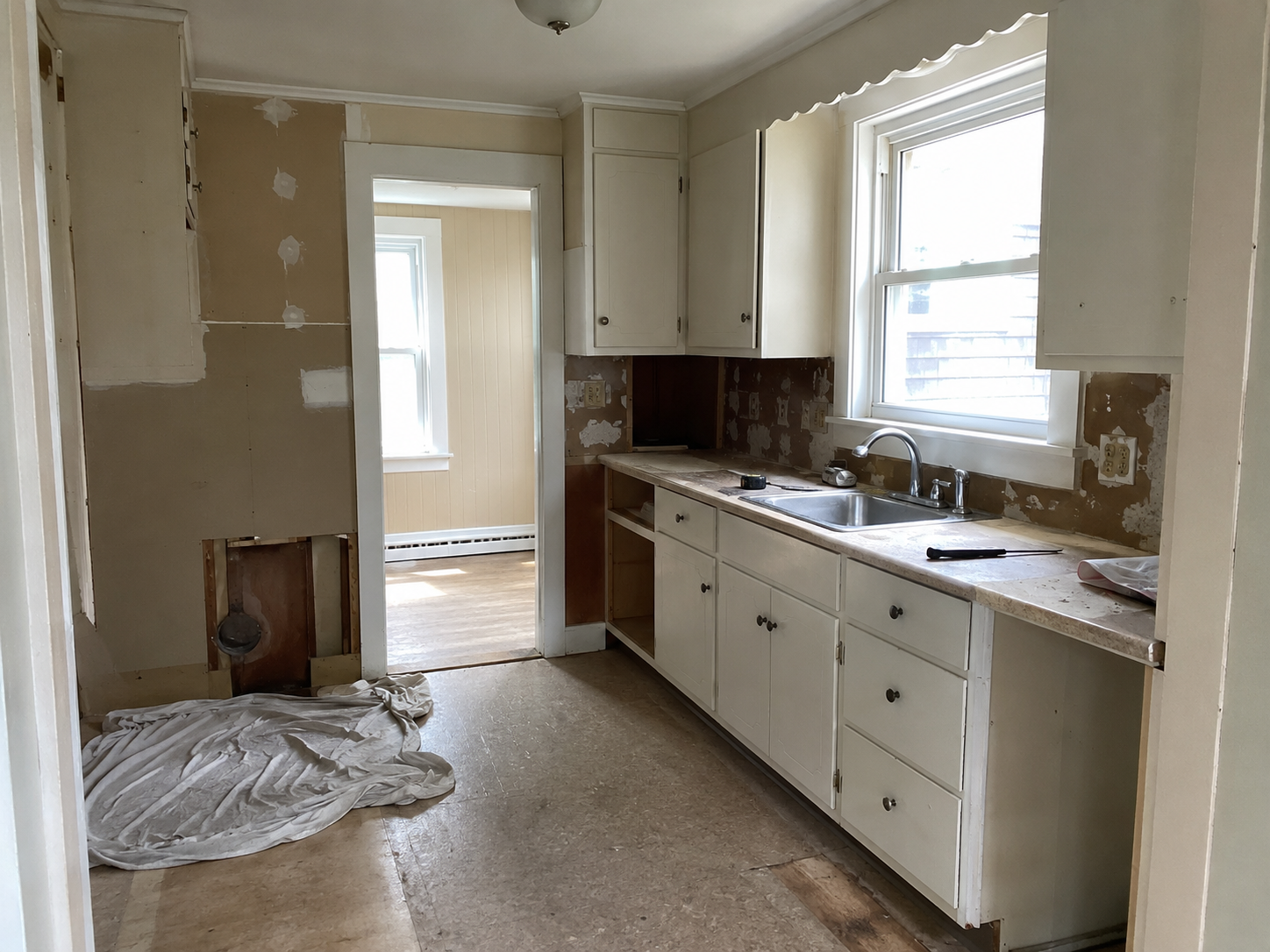 Small Cape Cod kitchen during remodeling with old cabinets, patched wall, window, and narrow layout.
