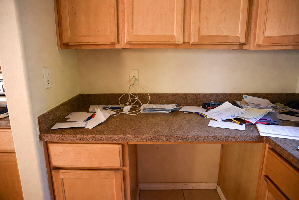 Early 2000s built-in kitchen desk with maple cabinets, granite counter, papers, and cords.