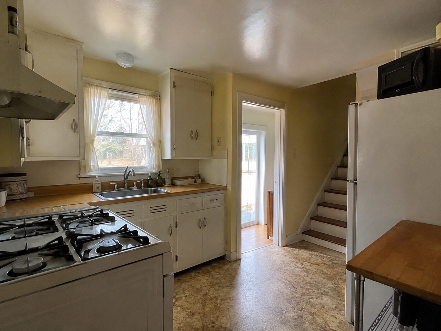 Small Cape Cod kitchen with sink window, stair, doorway, older cabinets, and tight layout.