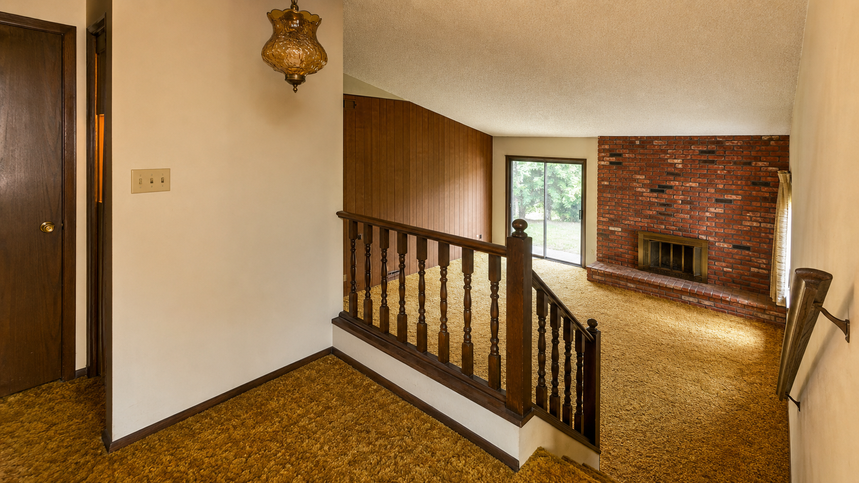 1970s split-level interior with wood railing, shag carpet, sliding door, and brick fireplace.