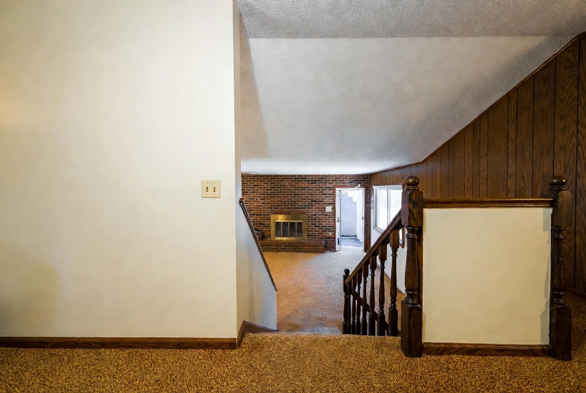 1970s split-level stair opening with shag carpet, railing, paneling, and brick fireplace below.