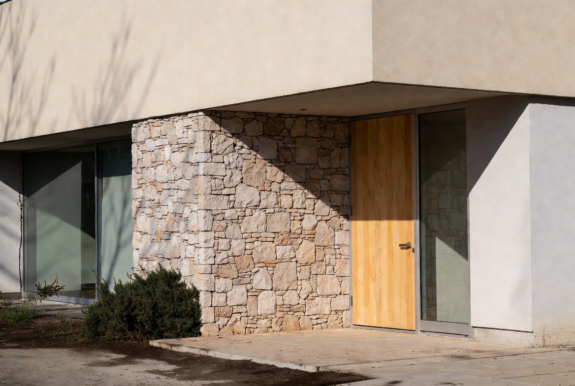 Modern entry with stone wall, stucco, wood door, glass, and concrete paving.